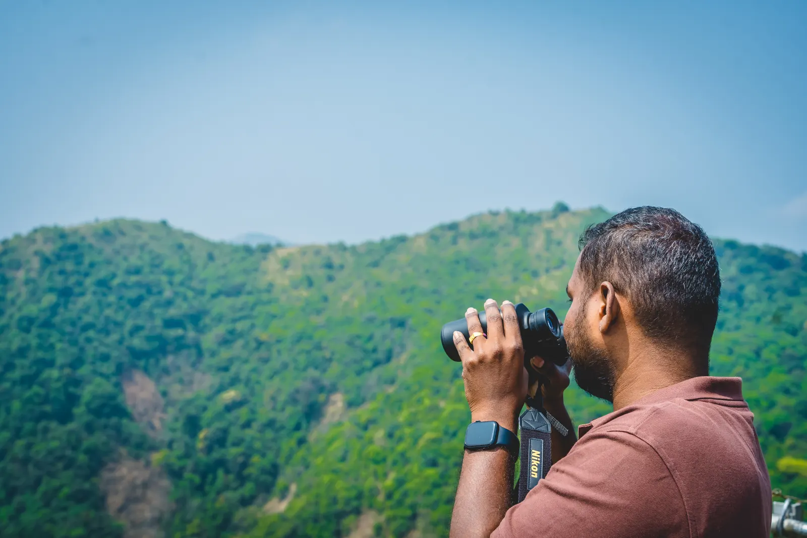A birder scanning a forested hillside through binoculars under a clear sky